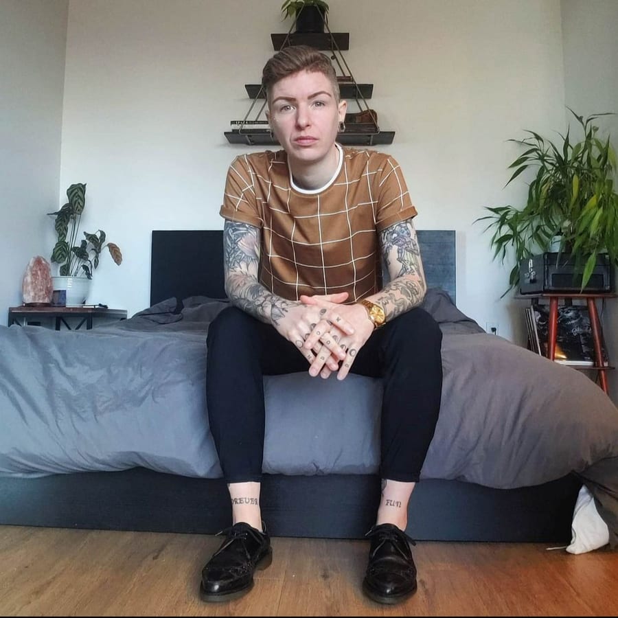 Jessi, the founder of Domino, sitting on the edge of a bed in a brown grid-print tee, black trousers, and polished black shoes